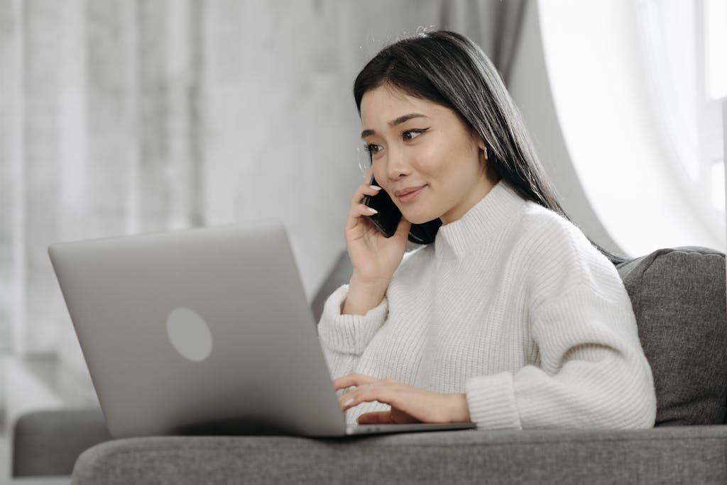 Woman multitasking from home using a laptop and smartphone on a sofa. Ideal for remote work themes.